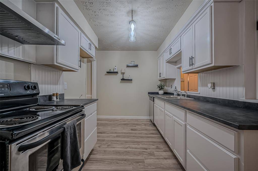 This is a galley-style kitchen featuring white cabinetry, dark countertops, with wood-look flooring. There's a textured ceiling and pendant lighting, offering a blend of traditional and contemporary elements.