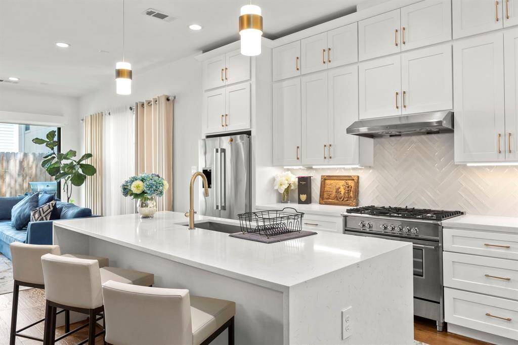 This gourmet kitchen is a chef's dream, featuring sleek stainless steel appliances, a large gas range, and a stylish herringbone tile backsplash. The expansive quartz island offers plenty of prep space, complemented by elegant gold fixtures and custom cabinetry for a clean, modern aesthetic.