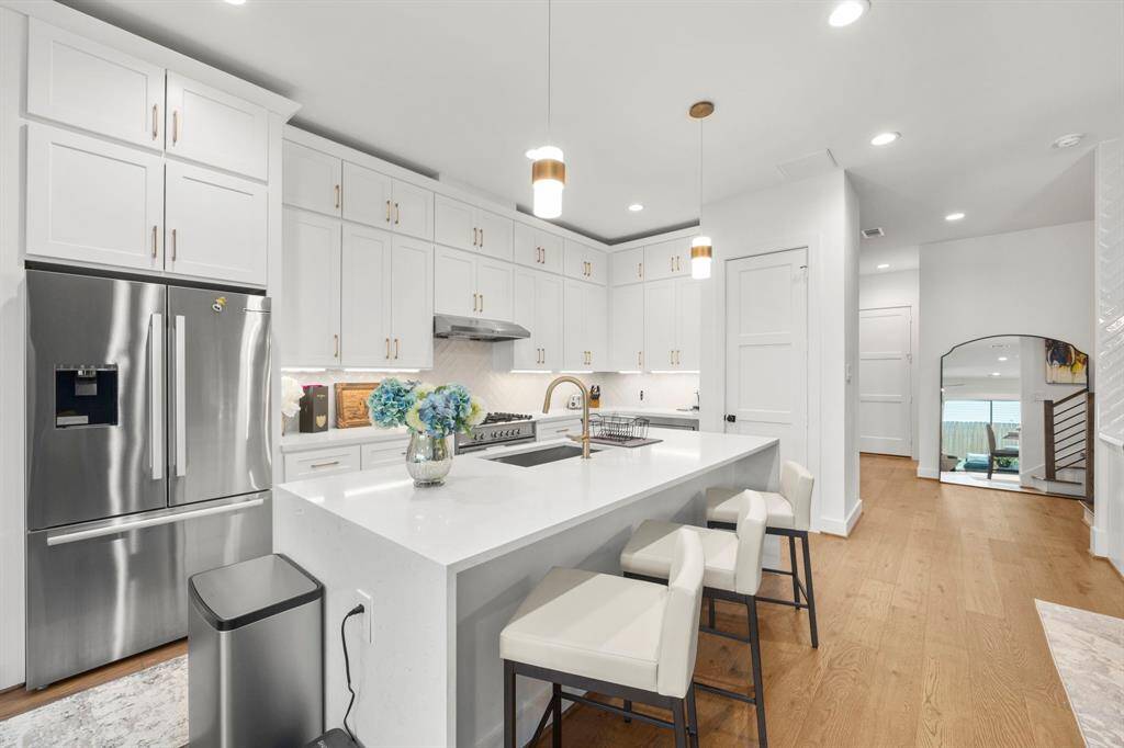 This sleek and spacious kitchen combines form and function with its large central island, modern stainless steel appliances, and ample storage provided by custom white cabinetry. Elegant pendant lights and warm wood flooring add a touch of luxury, while the open layout offers seamless flow into the adjoining living spaces, perfect for entertaining.