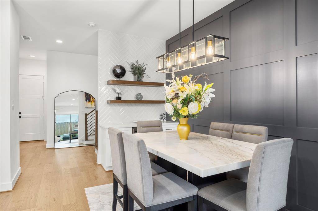 This dining room perfectly combines modern elegance with functional design. The sleek marble dining table is complemented by plush seating, while the bold accent wall and chic lighting fixture create a sophisticated atmosphere. The custom shelves and white herringbone backsplash in the bar area provide stylish storage and display space, adding to the room's overall appeal.
