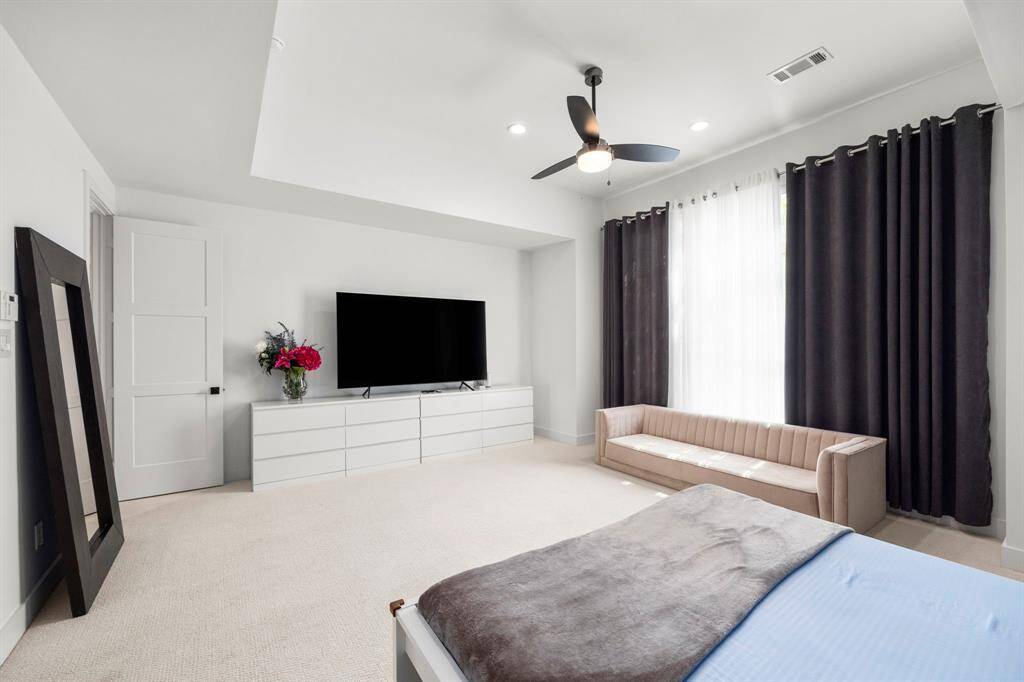 This spacious primary bedroom offers a serene retreat, featuring large windows that provide natural light while maintaining privacy with elegant drapery. The neutral tones and recessed ceiling create a peaceful and modern atmosphere, perfect for relaxation and unwinding.