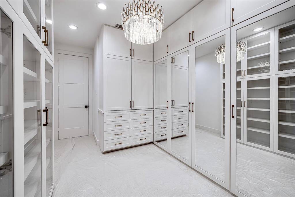 Additional view of the walk-in closet highlighting built-in shelving, sleek glass-front cabinets, and a stunning crystal chandelier that elevates the overall design. This closet provides both function and elegance with extensive storage options.