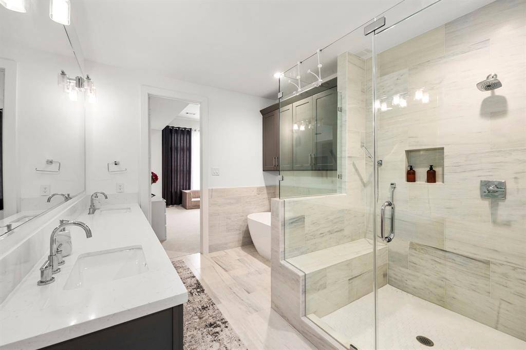 This primary ensuite bathroom offers a luxurious retreat, featuring dual sinks, a spacious glass-enclosed shower with built-in seating, and a deep soaking tub. The modern fixtures and clean lines create an elegant, spa-like atmosphere, perfectly complementing the serene aesthetic of the primary bedroom.