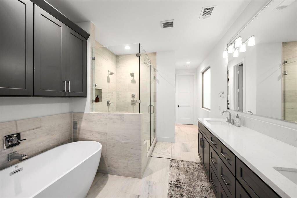 This additional view of the primary bathroom highlights the sleek freestanding tub and a spacious glass-enclosed shower with dual shower heads and built-in shelving. The large vanity offers ample storage space with modern dark cabinetry and polished finishes. The combination of clean lines, neutral tones, and luxurious fixtures adds a contemporary feel to the space.