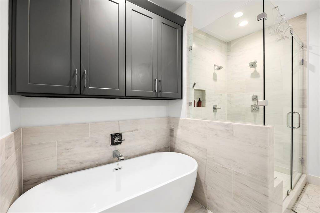 This close-up view of the primary bathroom highlights the freestanding soaking tub paired with sleek, modern fixtures. Above the tub, dark cabinetry provides extra storage space, complementing the neutral-toned tile work. Adjacent to the tub, the spacious glass-enclosed shower features dual shower heads and a built-in niche for added functionality. The clean lines and sophisticated design create a relaxing, spa-like atmosphere.