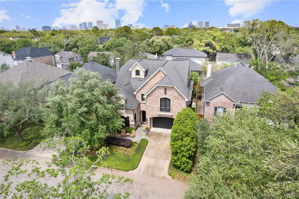 This photo showcases a beautiful two-story brick home nestled in a lush, tree-lined neighborhood. The property features a well-maintained front yard and a spacious driveway leading to a two-car garage. The surrounding area is dotted with similar homes, and the city skyline is visible in the background, indicating a convenient urban proximity.
