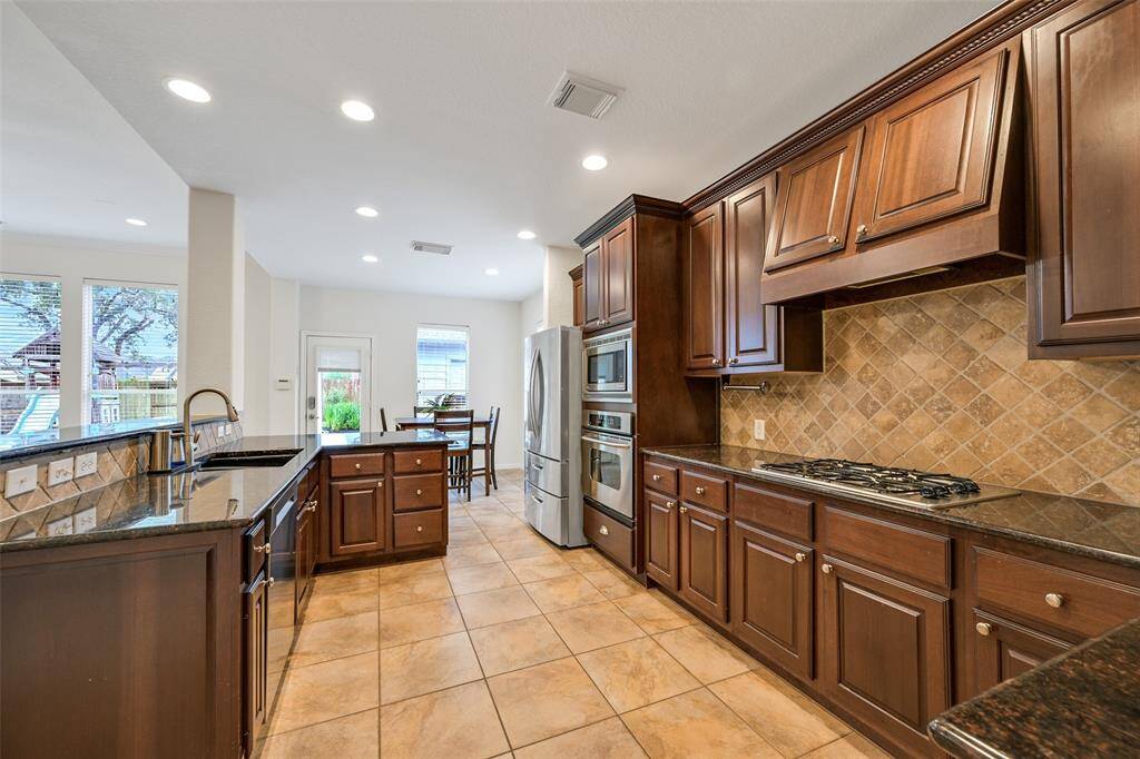 A wrap-around breakfast bar with granite countertops and ample seating space extends into this spacious kitchen with rich wood cabinetry, a diagonally laid tile backsplash, a gas cooktop, and double ovens—ideal for the home chef.