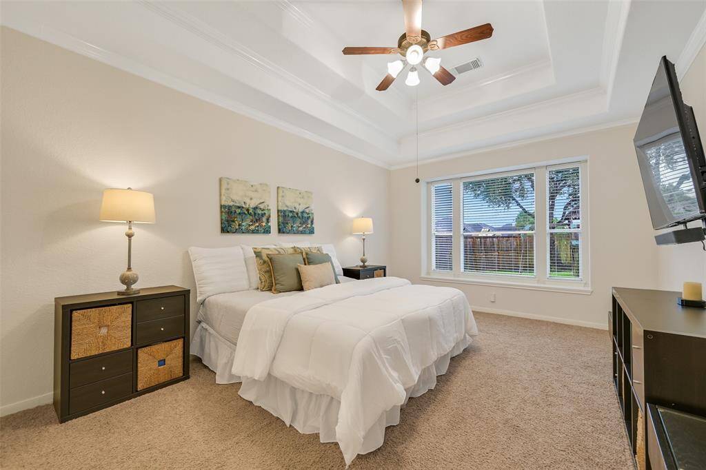 The first-floor primary suite features a double trayed ceiling trimmed with crown molding, soft neutral tones, separate his & hers closets, and a wall of windows overlooking the backyard for abundant natural light and a relaxing retreat.