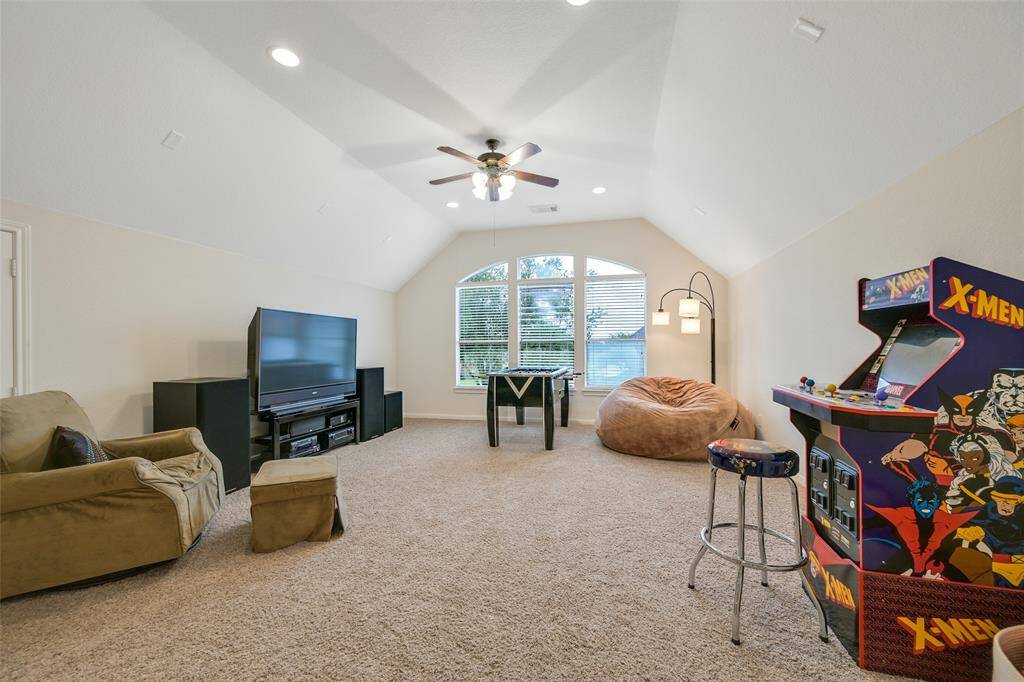 An alternate view of the spacious upstairs game room highlights the raised gambrel ceiling with recessed lights and a lighted ceiling fan, an oversized window with backyard views, and plenty of room for media, gaming, and lounge furniture.