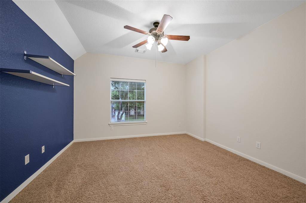 The fifth and final bedroom in the home features a raised gambrel ceiling with a lighted ceiling fan, plush carpet, a front-facing window with 2-inch blinds, a bold navy accent wall with floating shelving, and a roomy walk-in closet.