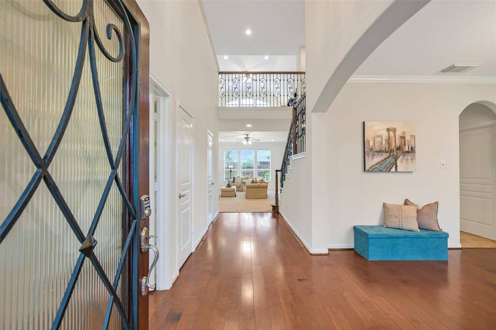 A decorative iron-accented front door with pleated glass and stained mahogany opens to a dramatic two-story foyer with a scrolled iron chandelier, engineered wood floors, fresh neutral paint, and a stunning wrought-iron split staircase.
