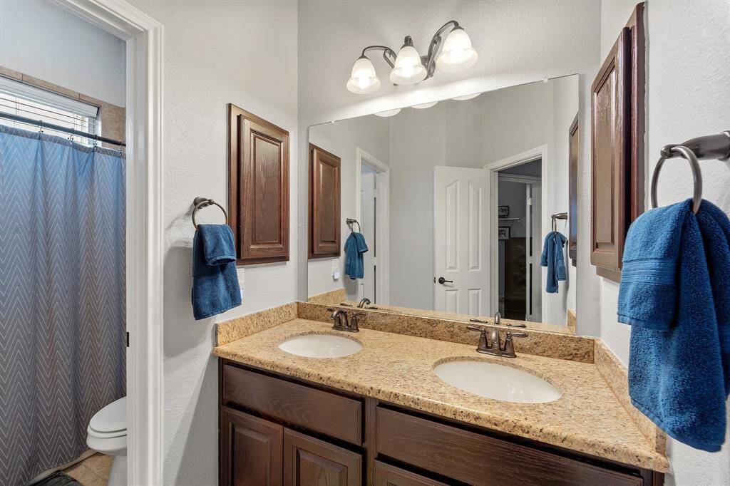 Secondary bathroom with granite countertops, a large mirror, double sinks, and dark stained cabinetry.