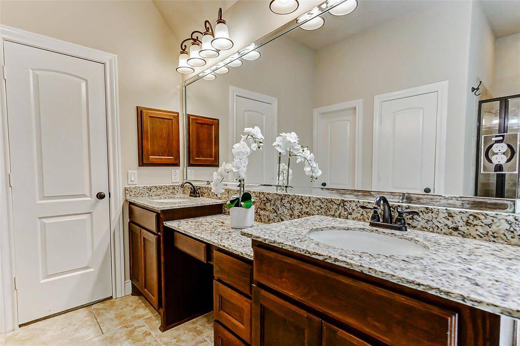 This spacious bathroom features a double vanity with granite countertops, elegant lighting, and a large soaking tub by a frosted window. The neutral tile flooring and high ceiling add a touch of luxury.