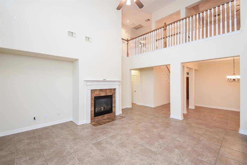 Spacious living area features high ceilings, a cozy fireplace, and a second-floor balcony overlooking the room. The neutral tile flooring complements the open layout, leading to a dining area with elegant lighting. Perfect for modern living and entertaining.