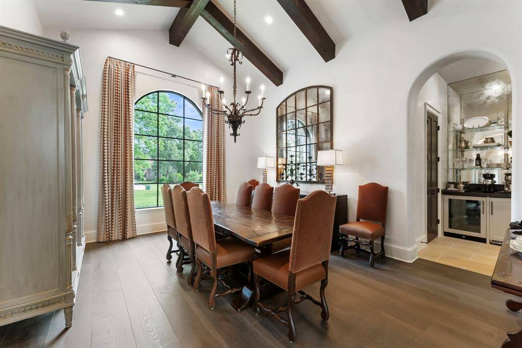 A sophisticated formal dining room, featuring warm wood floors, a vaulted ceiling with wooden beams, and beautiful windows that offer a picturesque view of the front exterior.