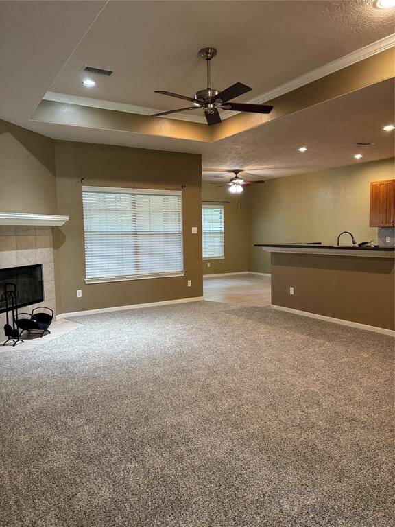 This inviting living space features plush carpet, a cozy fireplace, and an open layout leading to a kitchen area with a breakfast bar. Ceiling fans and recessed lighting add comfort and style.