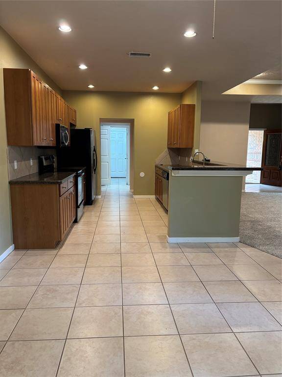 This photo showcases a modern kitchen with ample wooden cabinetry, stainless steel appliances, and a sleek countertop. The space is well-lit with recessed lighting and features a tiled floor, opening into a carpeted living area.