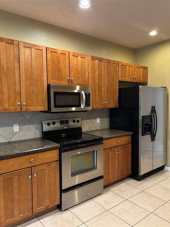 This kitchen features stainless steel appliances, including a stove, microwave, and refrigerator, complemented by wooden cabinets and granite countertops. The tile backsplash and flooring add a clean, modern touch, while recessed lighting provides ample illumination.