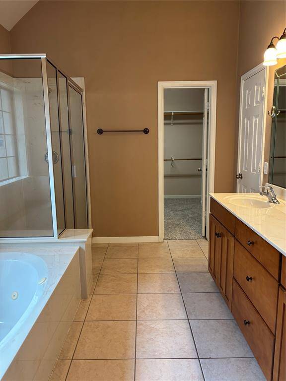 This spacious bathroom features a glass-enclosed shower, a large soaking tub, a double vanity with ample storage, and access to a walk-in closet. The neutral color palette and tile flooring add to the modern and clean aesthetic.