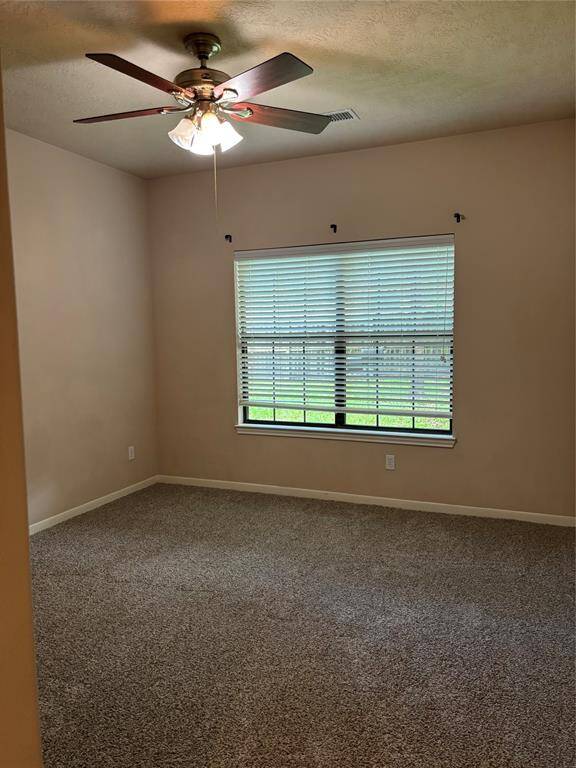 A cozy, carpeted room with neutral walls, a ceiling fan, and a large window with blinds, offering natural light and a view of the outdoors.