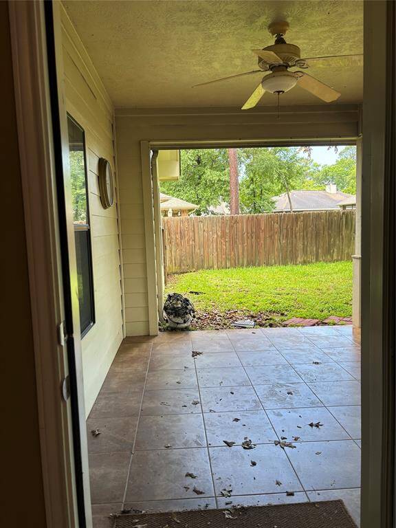 This photo shows a covered patio area with tiled flooring and a ceiling fan, leading to a grassy backyard enclosed by a wooden fence.