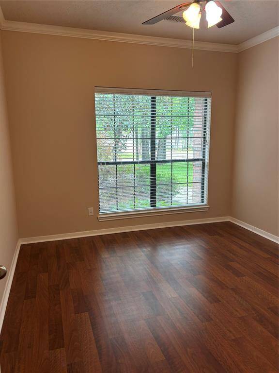 This room features warm wood flooring, a large window with blinds offering natural light and a view of the greenery outside. The neutral walls and ceiling fan with lights add to the cozy atmosphere.