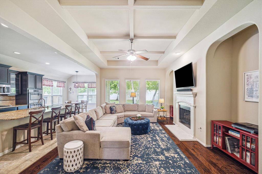 Step into this cozy family room, where warm wood flooring, soft neutral tones, and a raised coffered ceiling set the stage for comfort. A romantic gas-log fireplace adds an extra touch of charm, and a wall of windows frames views of the wooded backyard.