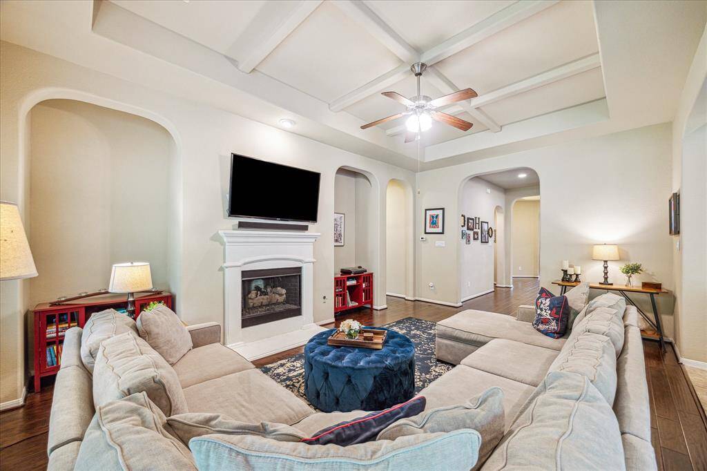 This family room is rich in architectural charm, featuring a striking coffered ceiling, graceful arched doorways, and alcoves flanking the gas-log fireplace to create a beautifully balanced focal point. Warm wood flooring and an open, airy layout make the space as inviting as it is elegant.