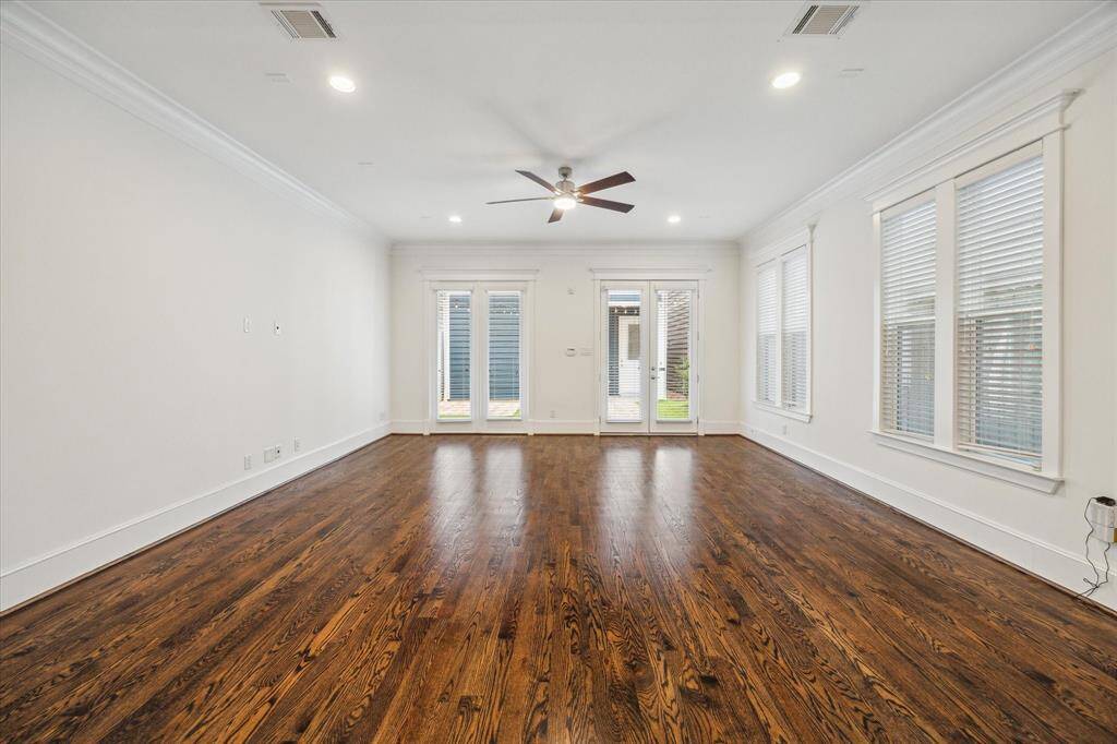 This expansive living area is designed for both comfort and style, featuring rich hardwood floors, crisp crown molding, and an abundance of natural light. Large windows along the side wall enhance the open and airy feel, while French doors lead to the backyard, creating a seamless transition for indoor-outdoor living. Recessed lighting and a modern ceiling fan add functionality, while the open layout allows for flexible furniture arrangements to suit your lifestyle.