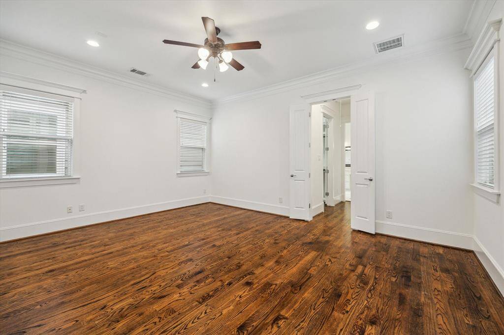 An inviting primary retreat blends timeless craftsmanship with modern luxury. Rich hardwood floors, crisp crown molding, and recessed lighting create a sophisticated yet cozy atmosphere. Large windows allow natural light to fill the space, enhancing the room’s warm and airy feel. Double doors lead to a spa-inspired ensuite bath, offering a seamless connection between relaxation and function.