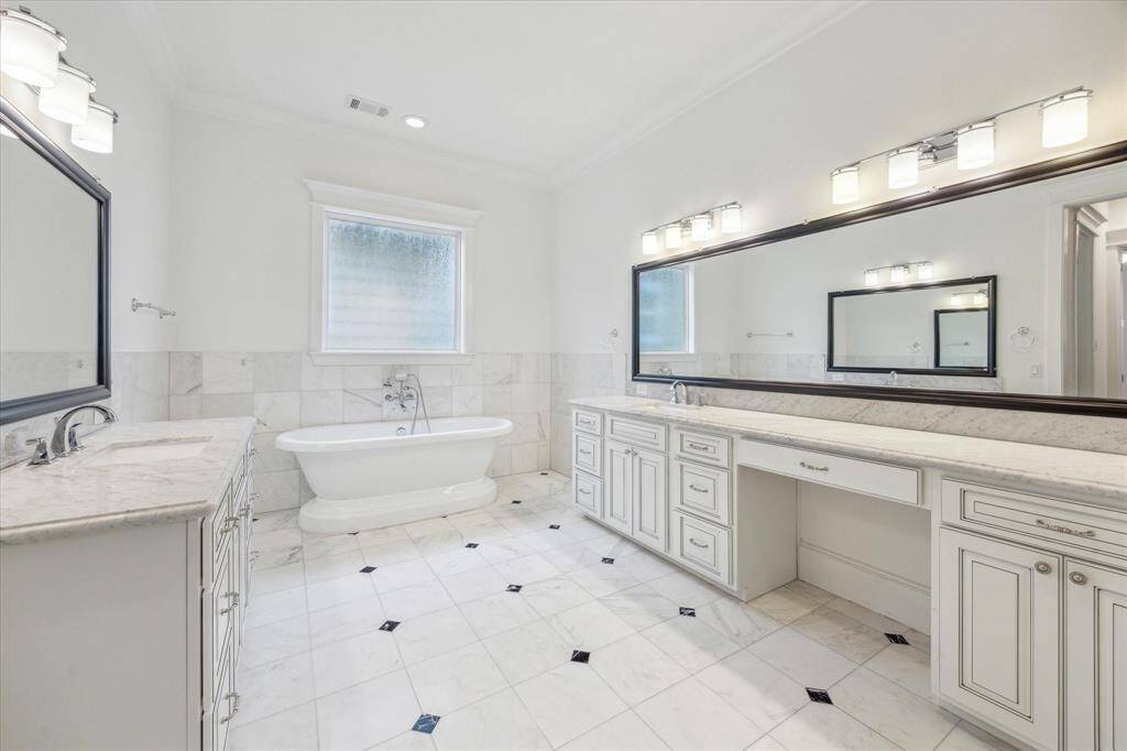 Luxury and elegance define this expansive primary bathroom, featuring classic tile flooring with elegant inlays, a freestanding soaking tub, and custom dual vanities with extensive counter space. The soft white cabinetry, accented by detailed trim and polished hardware, offers ample storage while maintaining a refined aesthetic.
