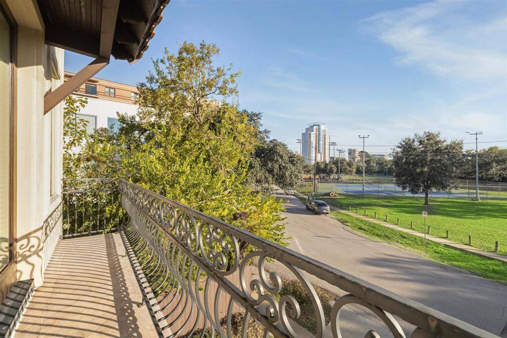 Charming balcony with ornate railing, overlooking a tree-lined street with a view of tennis courts and a city skyline in the background. It's a perfect spot for enjoying the outdoors and city views.