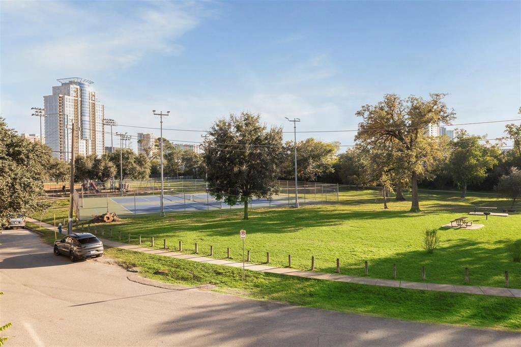 View from the balcony showcasing serene park with a tennis court, playground, and picnic area, surrounded by lush greenery. View of city skyline in the background, offering a blend of nature and urban living.
