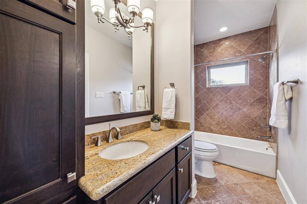 Secondary Hollywood bathroom features a granite countertop with a sink, elegant dark cabinetry, and a large mirror. The shower-tub combo is accented with stylish tile work, and there's ample lighting from a chic vanity light and recessed lights.