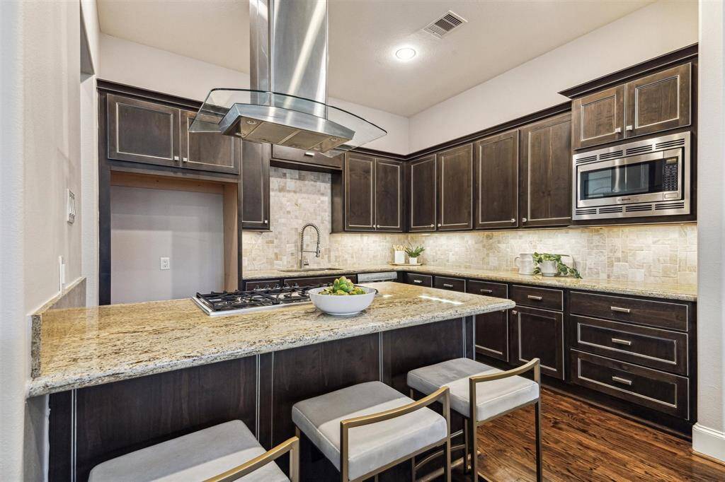 The kitchen features dark wood cabinetry, granite countertops, and a central island with a built-in gas cooktop and modern vent hood. It includes stainless steel appliances, under-cabinet lighting, and bar seating, creating a stylish and functional space.