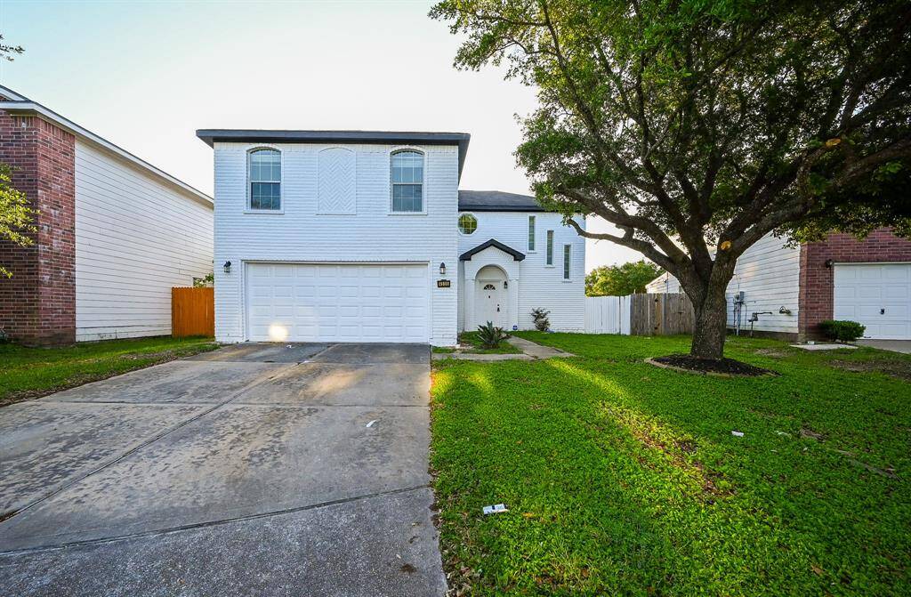This two-story, white brick home features a double garage and a spacious driveway. It has a charming arched entryway and a well-maintained front yard with a large, mature tree providing ample shade.