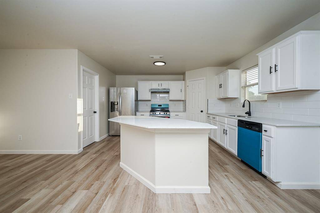 This modern kitchen features a spacious layout with light wood flooring, white cabinets, and stainless steel appliances. A central island provides additional workspace, and the room is brightened by natural light from a large window.