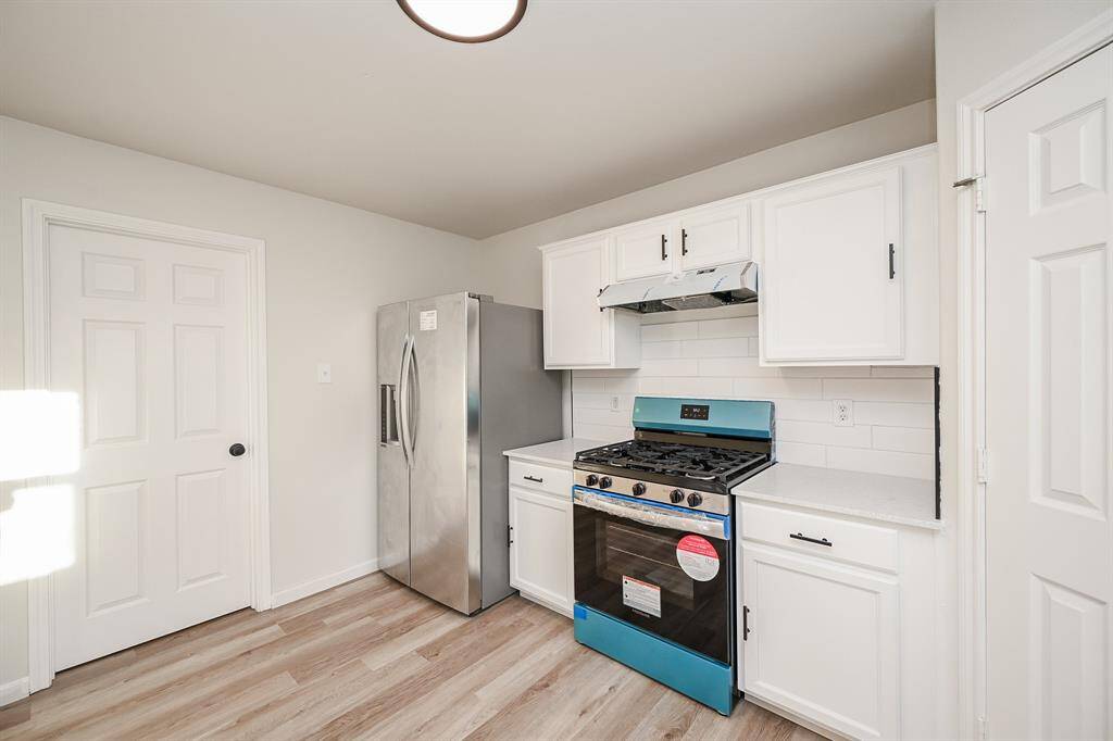 This kitchen features a modern design with a stainless steel fridge and gas stove, white cabinetry, and light wood flooring. Ample storage space and a clean, bright appearance make it inviting.