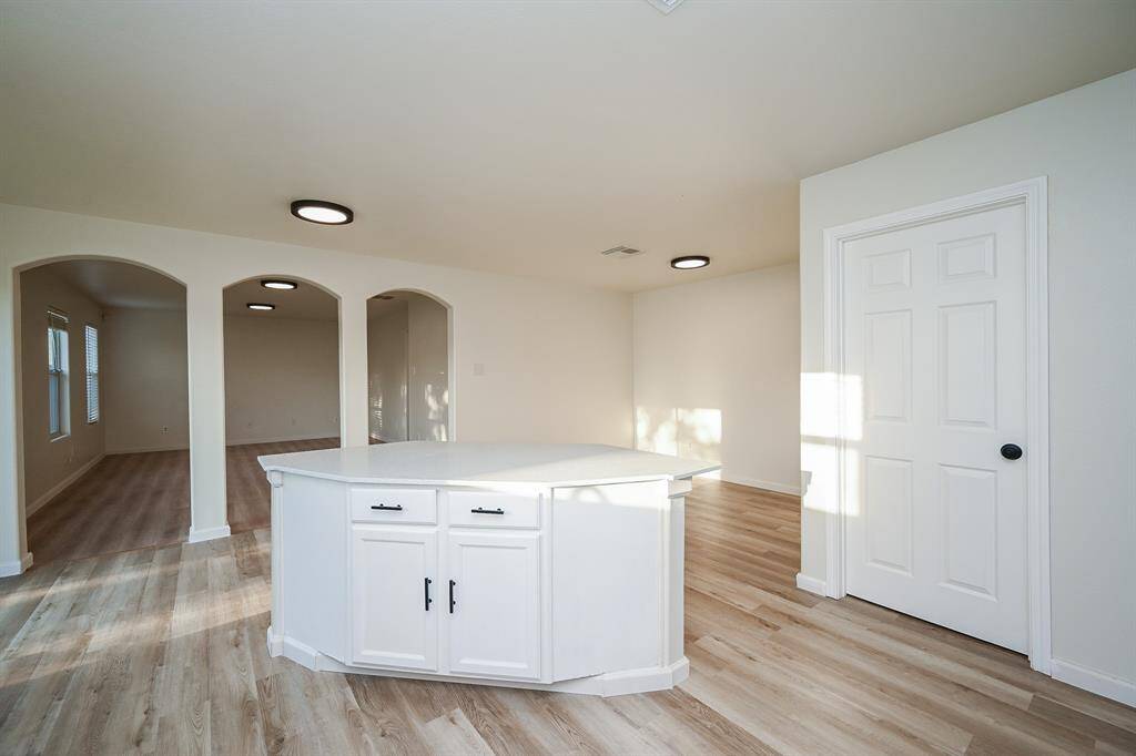 This spacious room features light wood flooring, neutral walls, and modern ceiling lights. It includes a central white cabinet, arched doorways leading to adjacent rooms, and ample natural light from large windows.