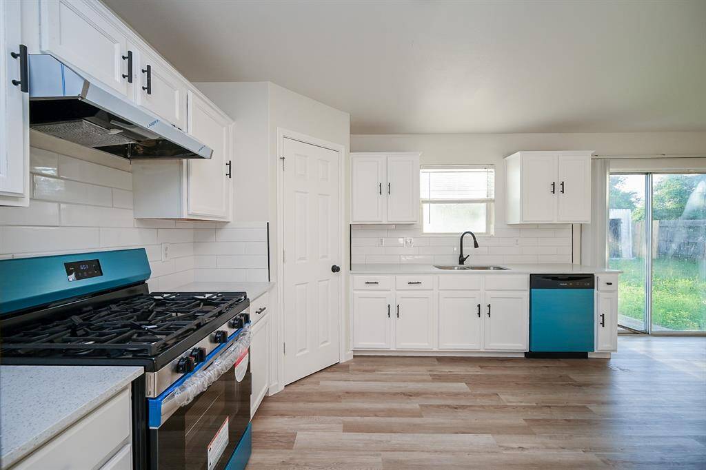 This kitchen features modern white cabinetry, a sleek stainless steel gas stove, and a brand new dishwasher. It has light wood flooring, ample natural light from a window and sliding glass door, and a spacious layout perfect for cooking and entertaining.
