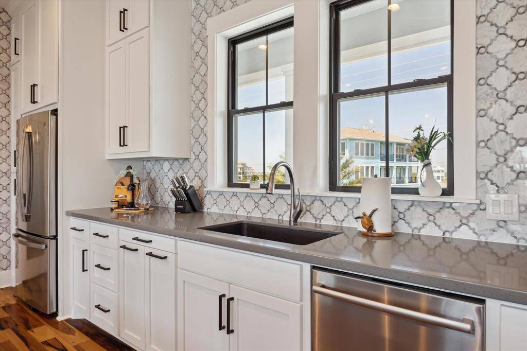 Modern kitchen with a clean and coastal aesthetic. The space features white cabinetry sleek quartz countertop, and a striking backsplash with an elegant arabesque tile pattern. Centered under two large windows is a deep, under-mount oversized granite sink with a contemporary faucet. The windows bring in natural light and offer waterfront views