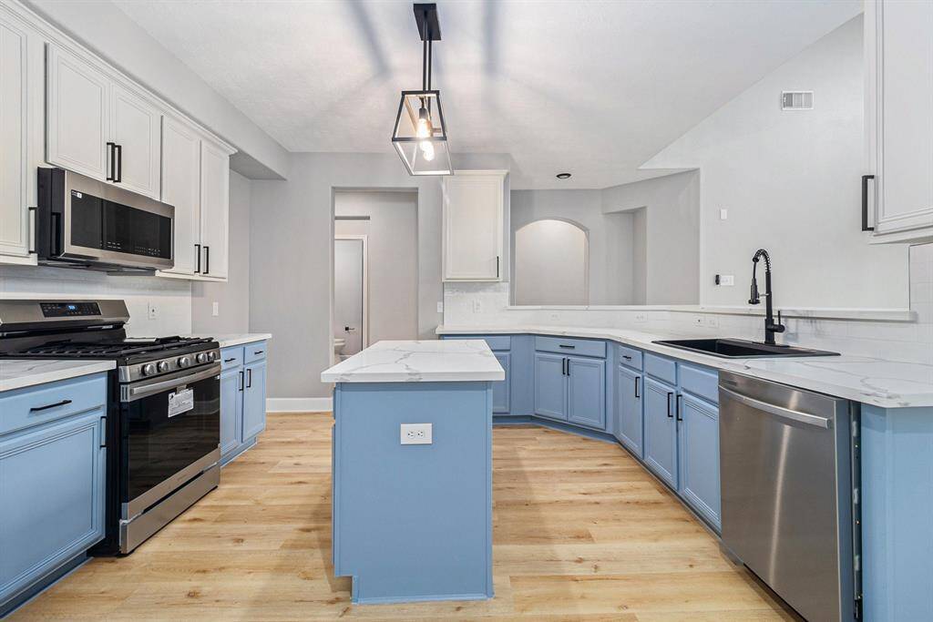 This kitchen boasts a modern design with light blue lower cabinets and white upper cabinets, complemented by stainless steel appliances. It features a central island with new countertop, a sleek black faucet, and new flooring for a welcoming atmosphere.