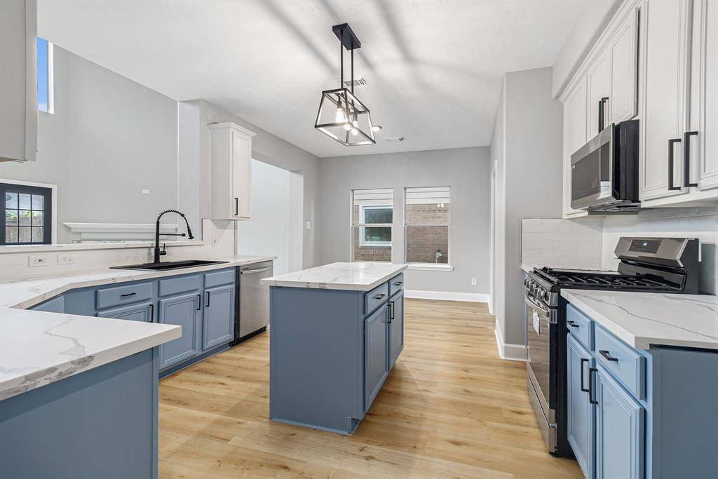 Modern kitchen with sleek blue cabinets, white countertops, and stainless steel appliances. Features an island, ample natural light, and new flooring.
