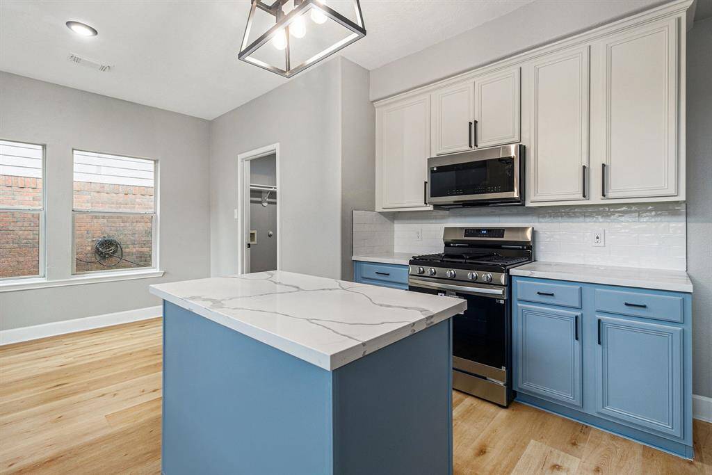 This modern kitchen features a stylish island with a marble-look countertop, blue lower cabinets, and white upper cabinets. It includes stainless steel appliances, a gas stove, and ample natural light from two large windows.