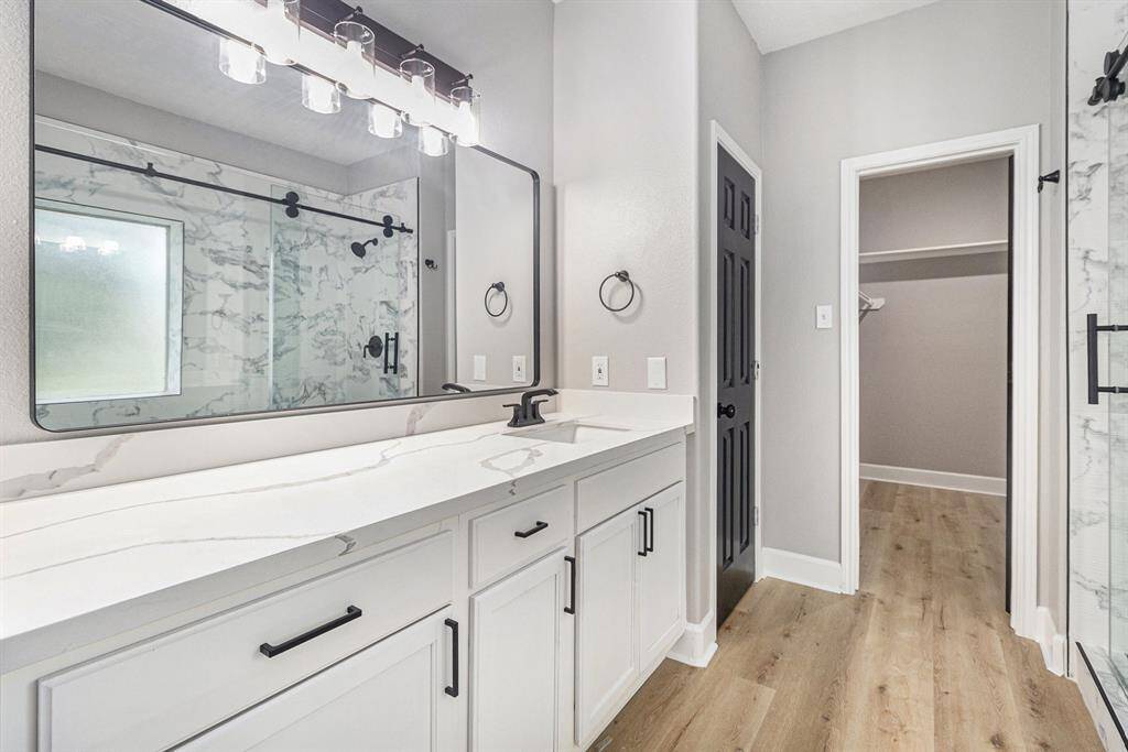 This photo showcases a modern bathroom featuring a sleek countertop and large mirror with stylish lighting to enhance the space, while the glass-enclosed shower adds a luxurious touch. A doorway leads to a spacious walk-in closet.