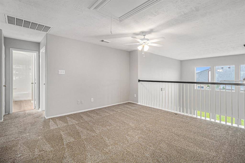 This upstairs photo showcases a bright, carpeted loft area with a ceiling fan and railing overlooking the floor below. It has access to a hallway leading to other rooms, offering a versatile space for a home office or play area.