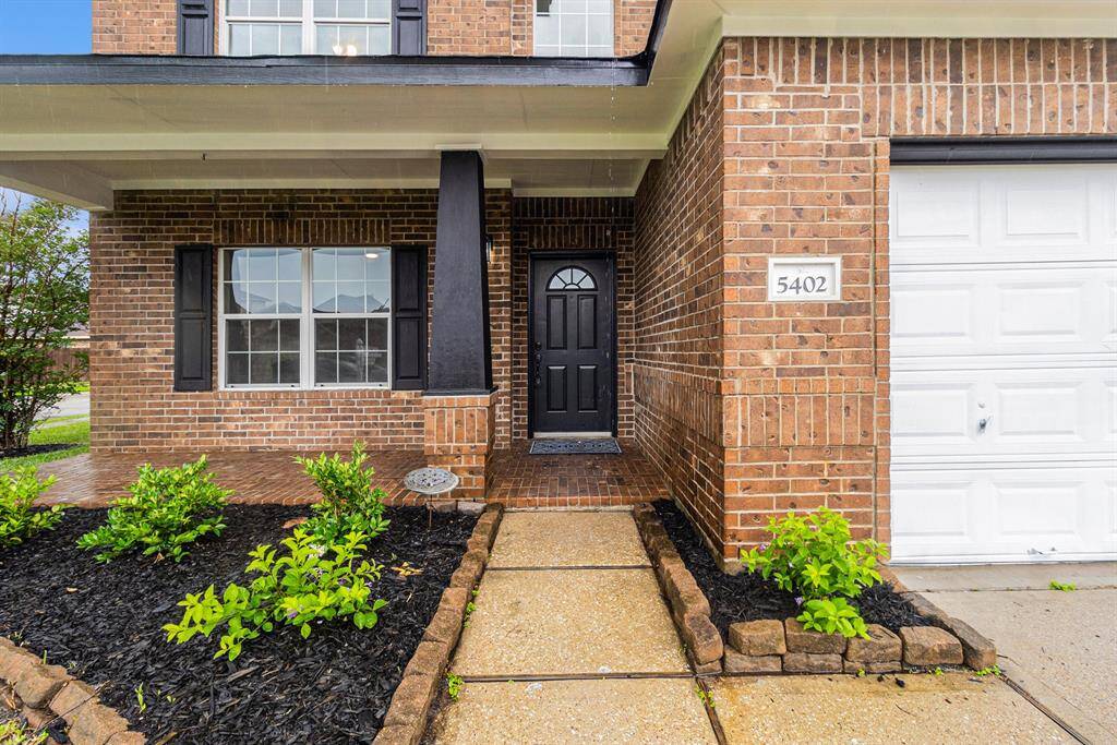 This photo showcases a welcoming brick exterior with a covered entryway, black front door, and well-maintained landscaping. The home features large windows with shutters and an attached garage.