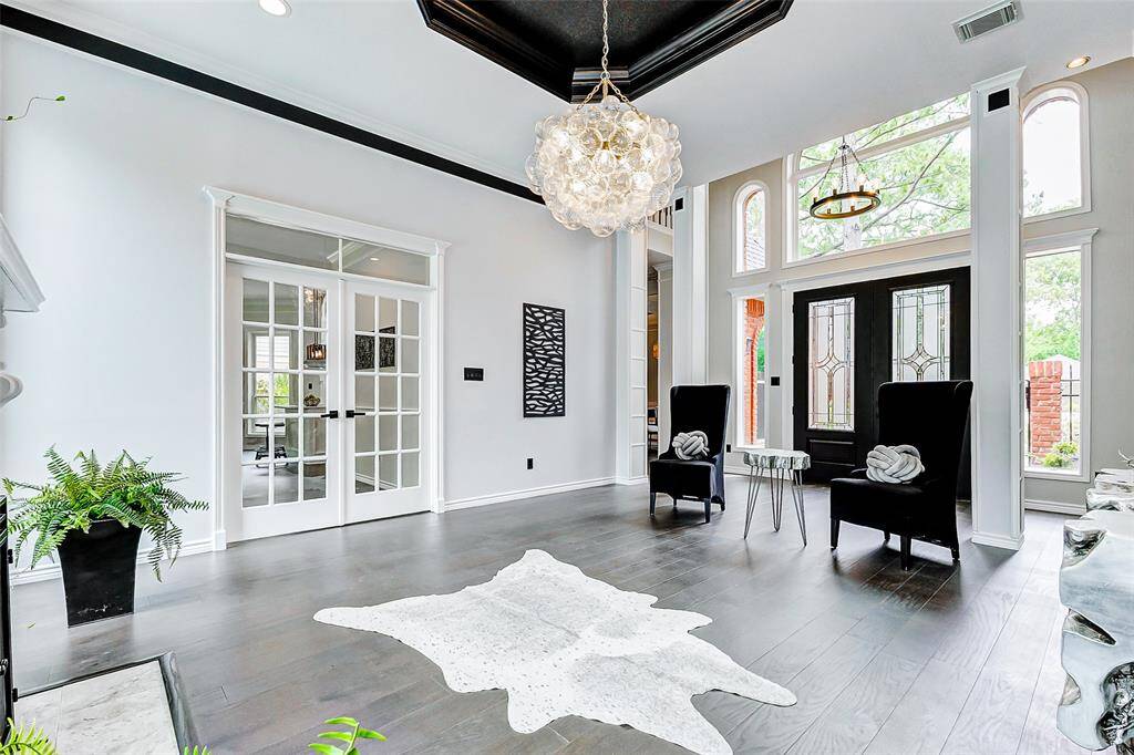 The elegant entryway features high ceilings and abundant natural light through large windows. It boasts a sophisticated chandelier, modern decor, dark wood flooring, and double doors leading to another room, creating an inviting and spacious atmosphere.