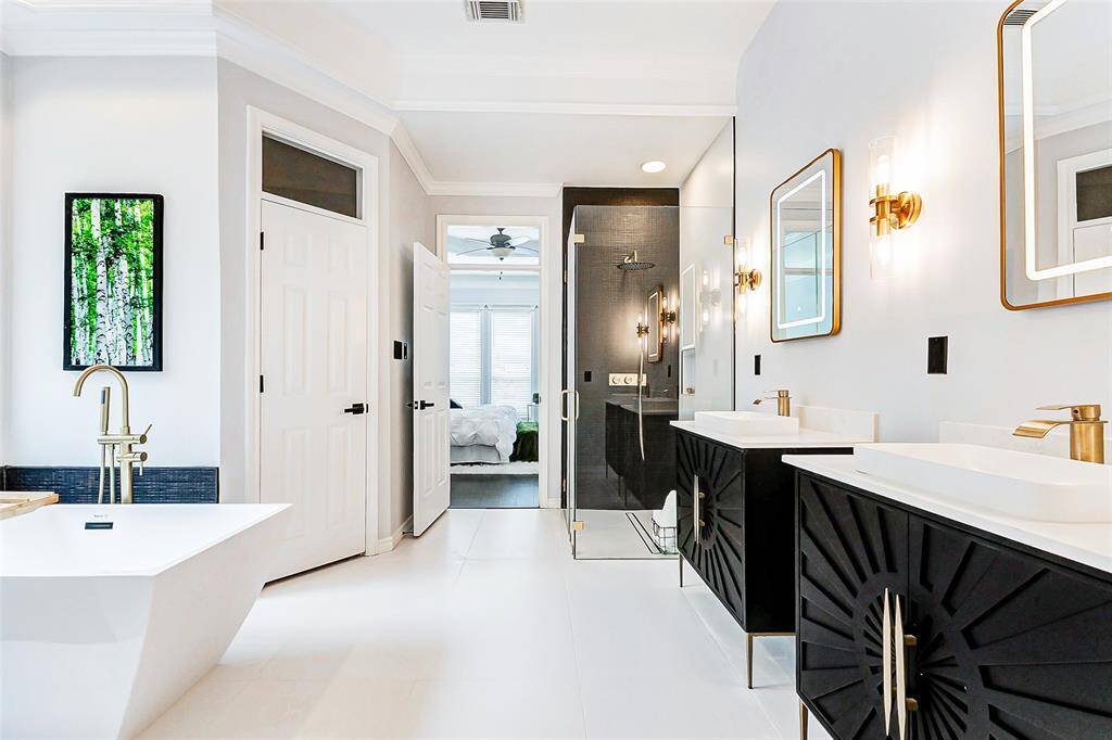 This modern bathroom presents a spacious layout with dual sinks, elegant black cabinetry, and gold fixtures. It includes a sleek glass-enclosed shower and a freestanding tub. The room is bright and airy, complemented by stylish lighting and a view into a cozy bedroom.
