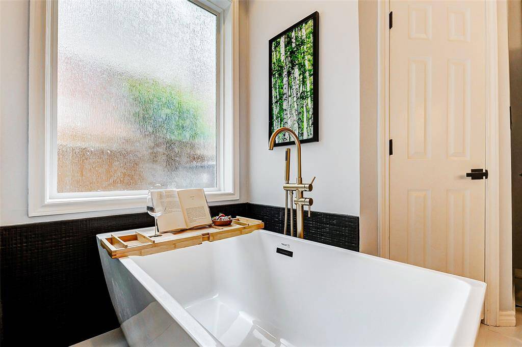 The stylish freestanding soaking tub with a sleek gold faucet is complemented by a large frosted window for privacy, offering a serene relaxation spot.